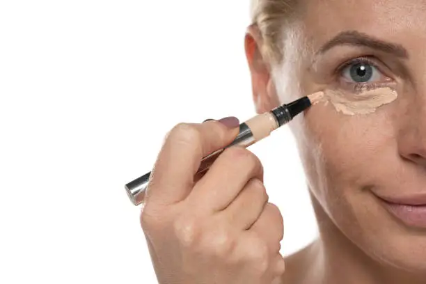 Photo of Middle aged senior woman applyes concealer under her eyes on white background