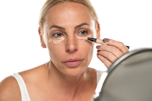 istock Middle aged senior woman applyes concealer under her eyes on white background 1581373374