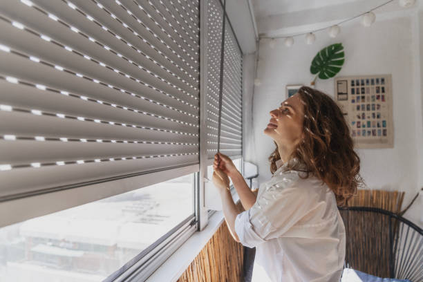 istock Young woman opening roller blind shutters on the balcony on a summer day 1581373404