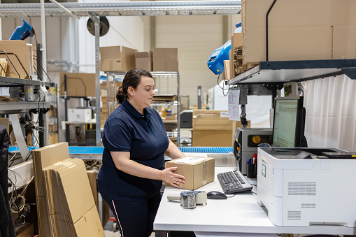 Side view of a woman working on computer and preparing a parcel for delivery in a distribution warehouse, Woman working in logistics center preparing shipment.