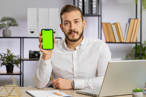 Young middle eastern developer business man hold smartphone with green screen chroma key mock up recommend good application promotional sale offer. Freelancer guy at office workplace looking at camera