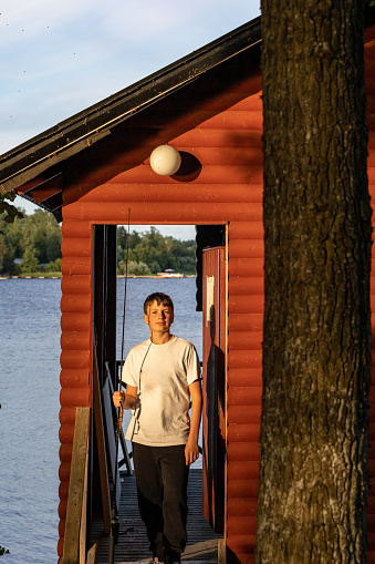 Stockholm, Sweden A 13-year-old boy with a fishing rod in front of a red cabin on Lake Malaren on a summer day poses in front of a door with a view over the lake.