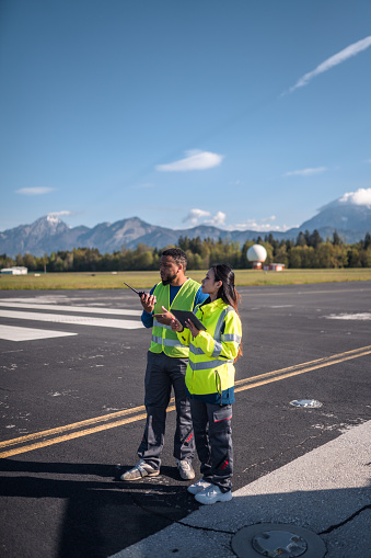 Airfield operations officers utilizing airfield landing and navigational aids. Wearing reflective clothes using a digital tablet and a walkie talkie to communicate with air traffic control.
