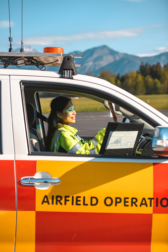 Asian female airfield operations officer sitting in a follow me car on an airport runway. She is wearing reflective clothes and ensuring safe take offs and landings.