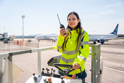 Asian female worker operating operating a container loader vehicle at the airport. She is wearing a reflective vest and using a walkie talkie.