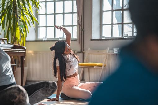 Mid adult Indian woman stretching to one side while doing a pilates workout. Other people in blurred foreground.