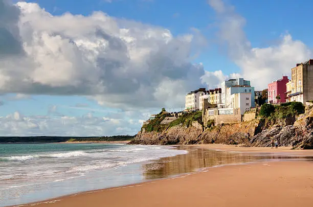 Pastel painted houses on the rocky coastline at Tenby, raised above Castle Beach, with the waves lapping on the sand, against a backdrop of blue sky with cumulus clouds.