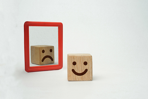 The concept of pretending to be happy but actually being sad. A wooden cube with happy emoticons but in mirror shows sad emoticons on white background