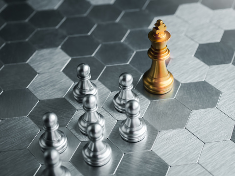 The golden king chess piece standing in front of silver pawn chess pieces on silver hexagon pattern board background. Leadership, follower, team, commander, competition, business strategy concept.
