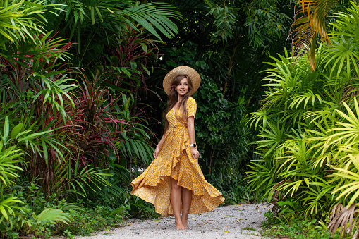 Female tourist girl with fashionable hat poses elegantly in lush green jungle. Young woman dancing charm and happiness in portrait of a tropical vacation.