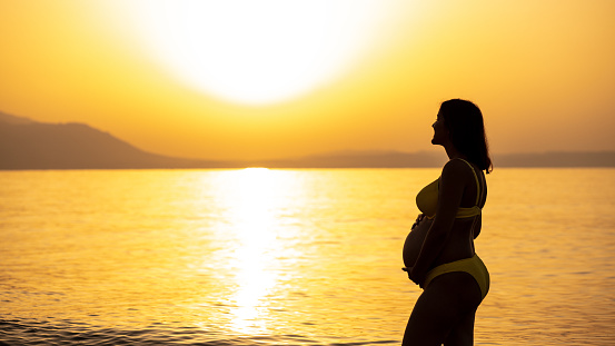 Photo of a eight month pregnant woman with hands on her abdomen at the seaside in sunrise