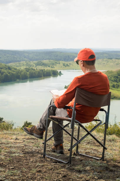 istock Man  sitting in camping chair outdoors and reading book. 1582193278