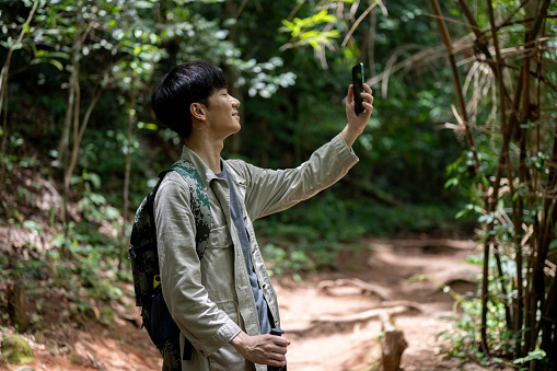 A handsome and happy Asian male hiker with a backpack and trekking gear is taking pictures with his smartphone while enjoying his hiking trip in the green forest.