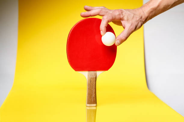 мужская рука, держащая мяч для пинг-понга над красной стороной ракетки для пинг-понга на желтом фоне. - racquette стоковые фото и изображения