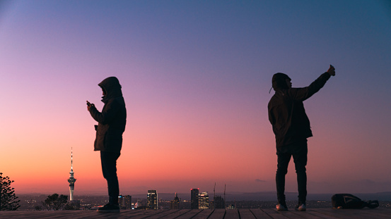 Two young man standing on viewing desk at Mt Eden enjoying city lights after sunset of Auckland Skyline and using smartphones.