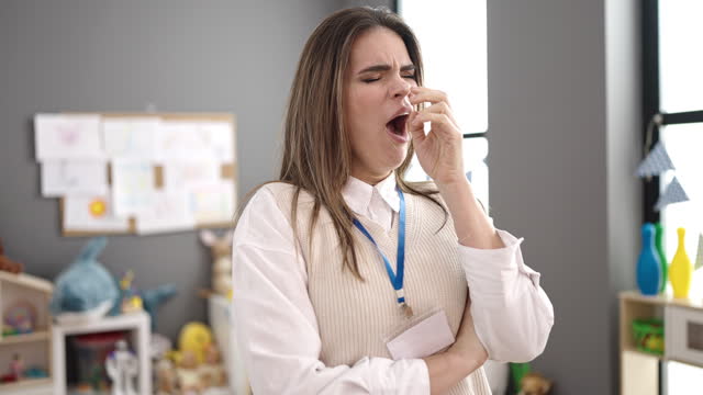 Young beautiful hispanic woman working as teacher yawning at kindergarten