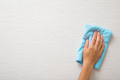 Young adult woman hand holding blue dry rag and wiping light gray wallpaper surface. Closeup. Front view. Empty place for text.