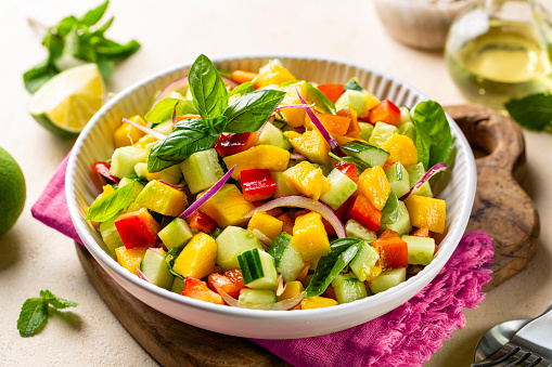 A bowl with healthy fresh mango salad made with red bell pepper, cucumber, red onion, fresh mint, basil and lime, olive oil and honey dressing.