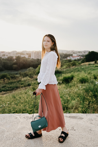 a girl with a green handbag in her hands walks near the park against the background of the city and the sunset. general plan