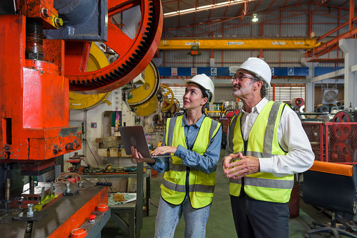 Two industrial workers engrossed in a discussion beside a high-tech assembly line machine, inside a bustling factory. Safety gear and intricate machinery enhance the sense of a reliable workflow.