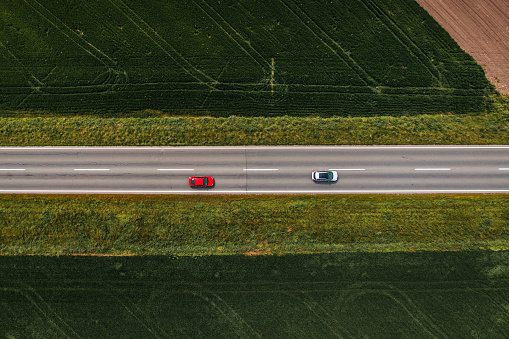 Two automobiles on straight asphalt highway through cultivated countryside landscape, aerial shot from drone pov, top view