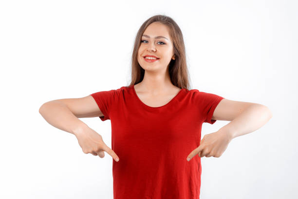 istock Smiling lovely girl points fingers down, shows advertisement on blank white space. Young brunette female looking happy by promotion offer, stands over white background 1582515434
