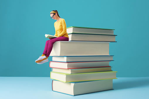 Tiny smart woman sitting on a pile of books and reading, education and learning concept