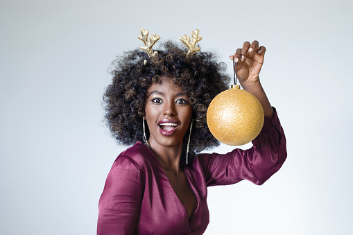 Portrait of happy young woman wearing maroon elegant dress and Christmas headband holding big xmas ball and laughing at camera. Studio shot, white background.