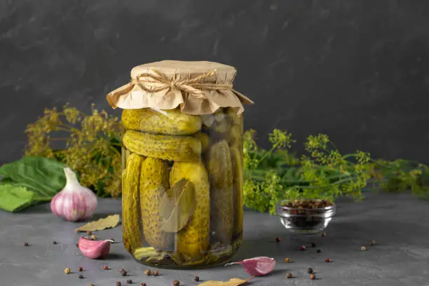 Homemade pickled cucumbers with garlic, horseradish, pepper and dill in glass jar on gray background