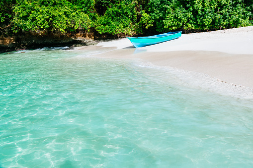 A blue boat on a white sand beach. The boat is small and wooden, with a white stripe on the side. The beach is white and sandy, with a green tree line in the background. The water is clear and turquoise, and is calm. The sky is not visible in the image. This photo can be used for travel, tourism, nautical, or coastal themes.