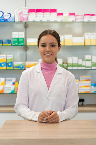 young female pharmacist looking at camera from pharmacy counter with products on shelves in the background