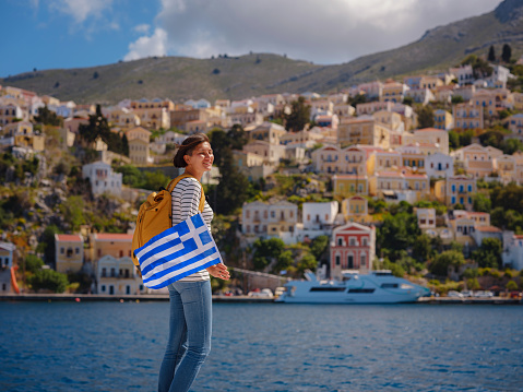 Nice asian Happy Female with backpack anf greek flag Enjoying her holidays on Symi Islands. View of port Symi or Simi, is tiny island of Dodecanese, Greece, calm atmosphere and fabulous architecture.