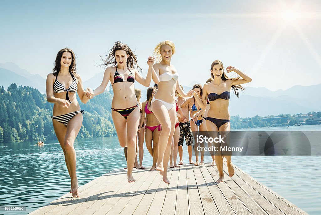 Cheering Girls at the Lake Summer Vacation Group of cheering young adults, girls in bikinis in the front row, running and jumping over a jetty at a lake on a beautiful sunny summer day. Bikini Stock Photo