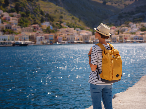 Nice asian Happy Female with backpack Enjoying her holidays on Symi Islands. View of port Symi or Simi, is tiny island of Dodecanese, Greece, calm atmosphere and fabulous architecture.