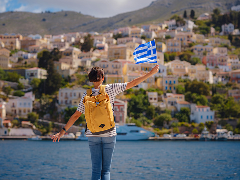 Nice asian Happy Female with backpack anf greek flag Enjoying her holidays on Symi Islands. View of port Symi or Simi, is tiny island of Dodecanese, Greece, calm atmosphere and fabulous architecture.