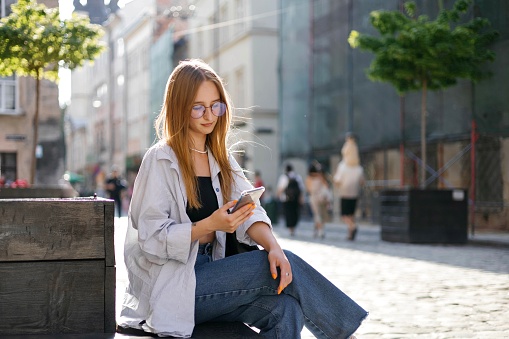 A young woman with a smartphone, wearing glasses, having bright nails, sitting on bench in the streets of Lviv during the summer