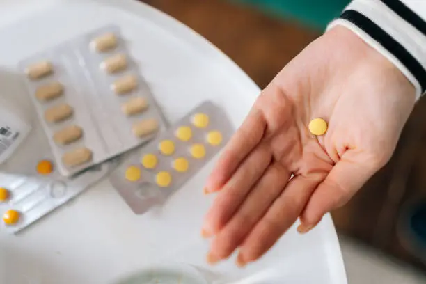 Close-up top view of unrecognizable sick young woman holding medicine for flu, influenza, painkiller, vitamin complex, oral contraceptives on hands on blurred background of table with various drugs.