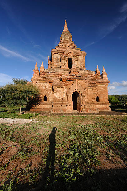 A pagoda in Bagan against blue sky, with a photographer/tourist's shadow foreground.