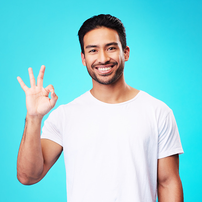 Happy, perfect gesture and portrait of a man in a studio with an agreement sign or expression. Happiness, smile and young Indian male model with an approval hand emoji isolated by a blue background.