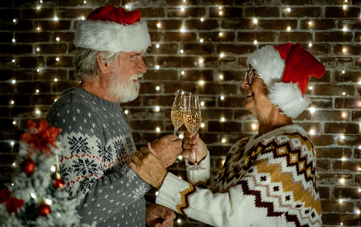 Christmas time. Happy senior family couple in Santa hat toasting looking in the eyes. Elderly man and woman celebrating Christmas at home drinking wine