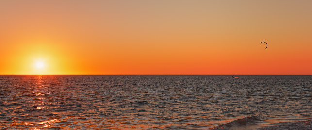 Sun setting down to calm sea, bright orange red sky, silhouette of kite surfer in distance
