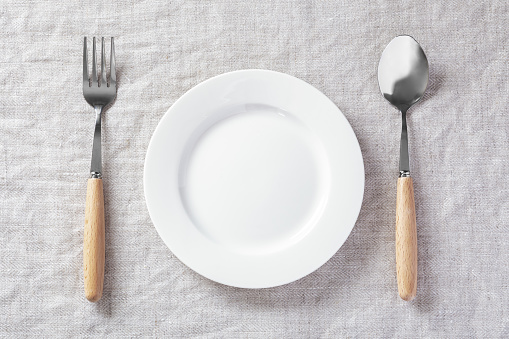 White plate with fork and spoon on linen tablecloth, table setting, top view