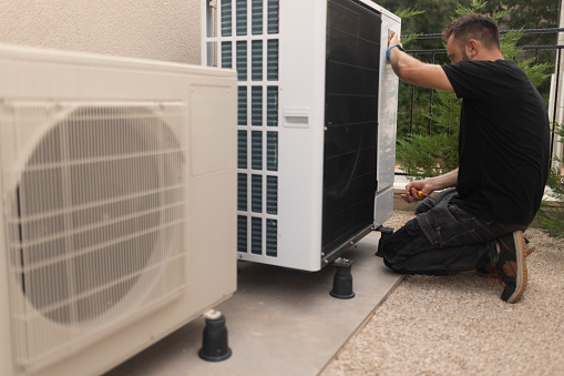 A male electrician is standing in the yard installing a heat pump
