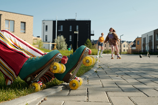 Lower part of legs of young active female teenager in roller skaters and pants sitting by trottoire and having rest against two girls skating