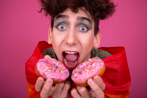istock Funny drag queen poses in the studio on a pink background and eats donuts. 1582947151