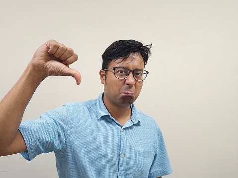 An Indian male wearing glasses and a blue shirt is expressing his dissatisfaction by making a thumb down gesture against a neutral gray background
