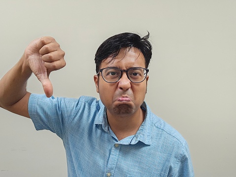 An Indian male wearing glasses and a blue shirt expressing his dissatisfaction by making a thumb down gesture against a gray background