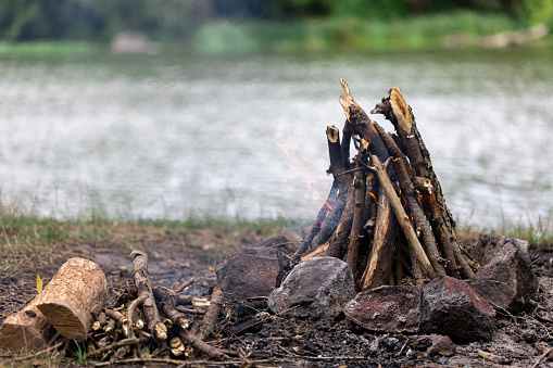 Close up of burning brushwood campfire on forest ground on blurred background of river, camp fire in cloudy day outdoors.