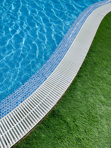 edge of an s shaped swimming pool, with crystal clear water, a white pool trellis and green grass, vertical