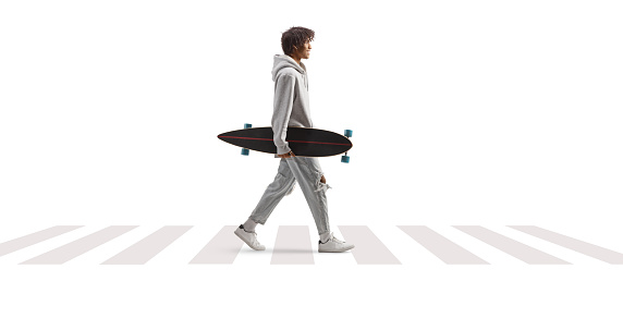 Full length profile shot of an african american guy carrying a longboard and walking at pedestrian crossing isolated on white background